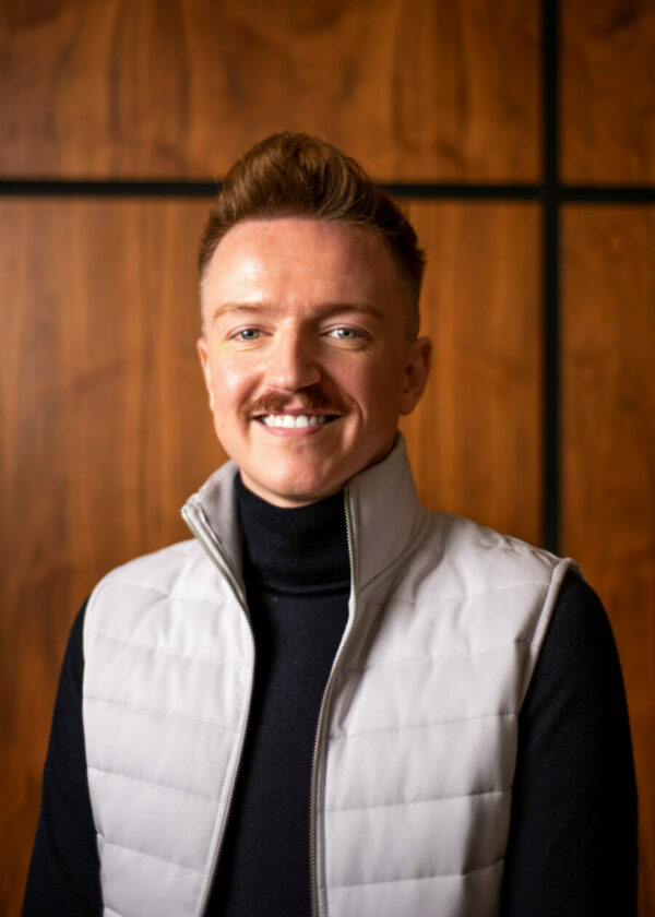 A portrait of a grinning man with auburn hair, a moustache, and a light gray vest, posing against a wooden background.