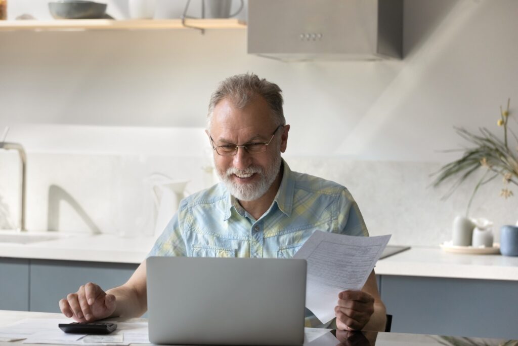 Smiling older man in glasses using a laptop and holding paperwork at a kitchen counter.